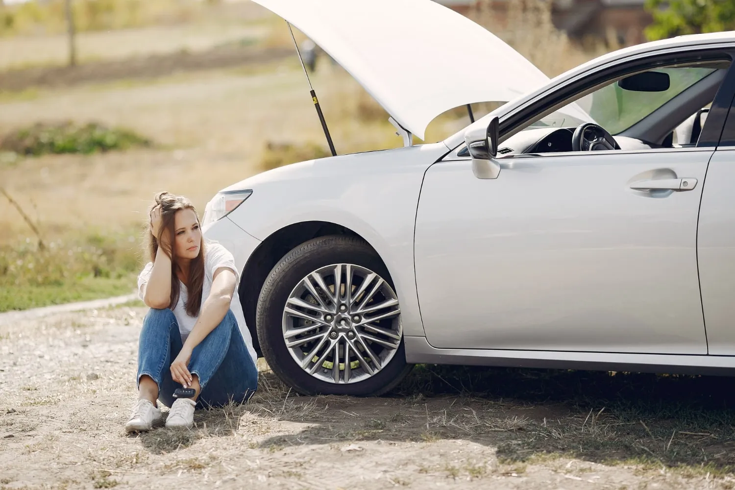Young woman sitting beside a white car with an open hood, appearing frustrated, on a sunny day