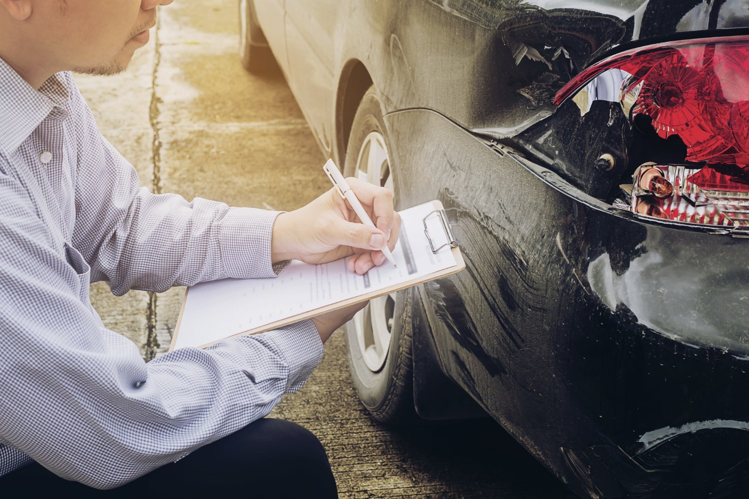 Insurance agent writing on clipboard after inspecting damage on a car's rear bumper post-collision
