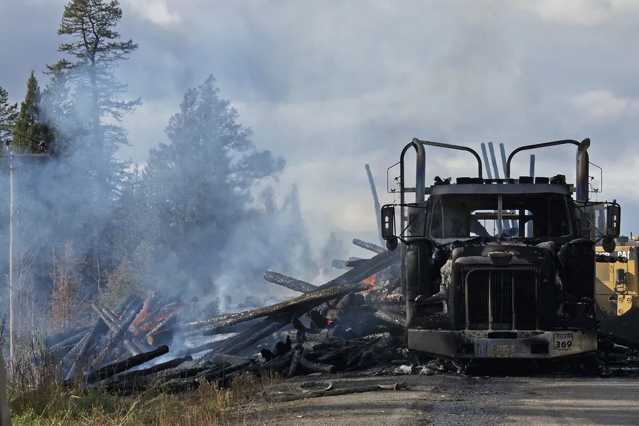 A charred truck surrounded by burnt logs and smoke after a severe accident