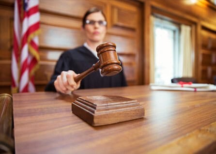 A judge in a courtroom is holding a gavel above a wooden desk with an American flag visible in the background.