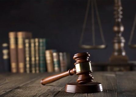 A wooden gavel rests on a table in front of law books and scales of justice in a dimly lit setting.