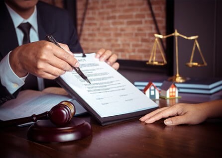 A person in a suit points at a contract with a pen. A gavel, miniature houses, and scales of justice are on the table.