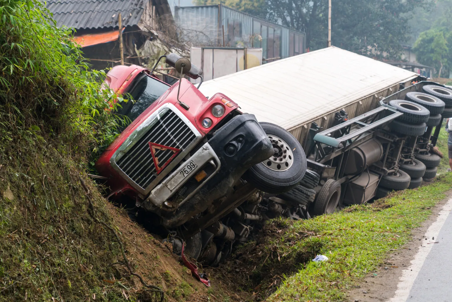 A large red truck has tipped over onto its side in a ditch beside a road, with its cab facing uphill and trailer off the ground.