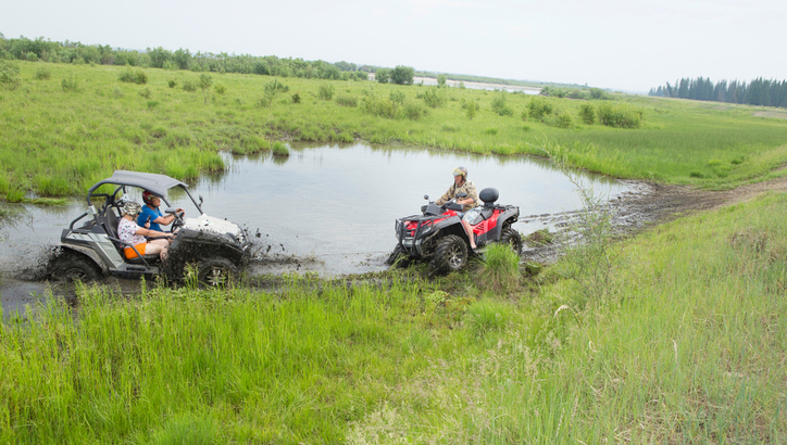 Two people in a white side-by-side off-road vehicle and one person on a red ATV drive through a muddy area in a grassy field.