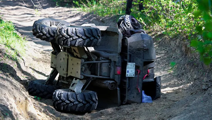 An ATV lies flipped on its side in a dirt path, with its undercarriage exposed and a person wearing a helmet partially visible behind the vehicle.