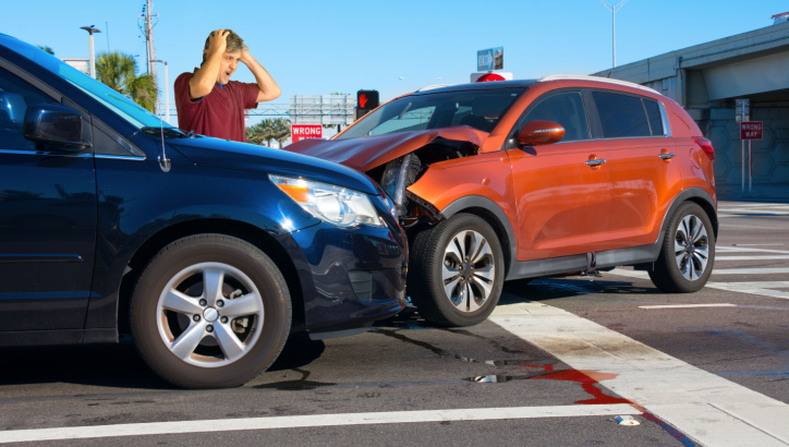 Two cars have collided at an intersection; one person stands nearby holding their head in distress as fluid leaks onto the road.