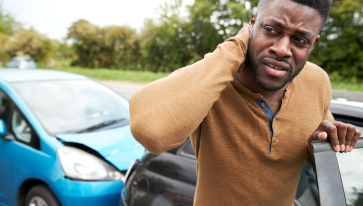 A man stands outside his car holding his neck, appearing in pain, after a rear-end collision with a blue car on a road.