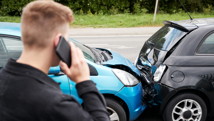 A man on the phone stands near two cars involved in a rear-end collision on the side of a road.