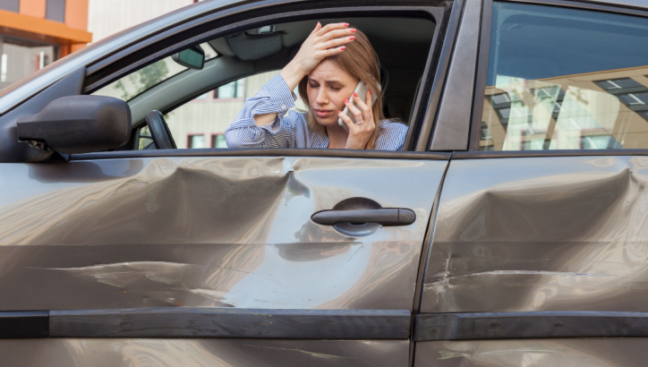 A woman sitting in a car with a dented door looks distressed while talking on her phone, possibly reporting the accident.