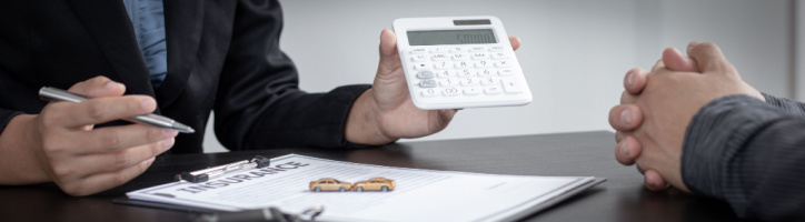 Two people sit at a desk discussing insurance; one holds a pen near an insurance form and toy cars, the other holds a calculator.