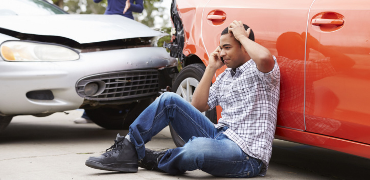 A man sits on the ground next to an orange car, holding his head and talking on the phone after a collision with a silver car.