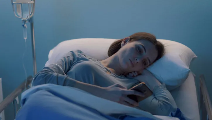 A woman lies in a hospital bed under a blue blanket, holding a smartphone and looking at the screen. An IV drip is visible beside the bed.