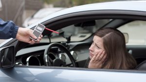 A woman in a car looks at a breathalyzer device held by a person outside the driver's window.