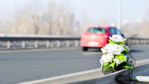 A bouquet of white artificial flowers is attached to a highway guardrail, with a red car driving away in the background.