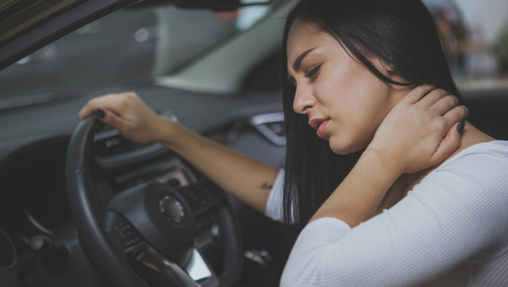 A woman sits in a car driver's seat, holding her neck with one hand and appearing to be in pain or discomfort.
