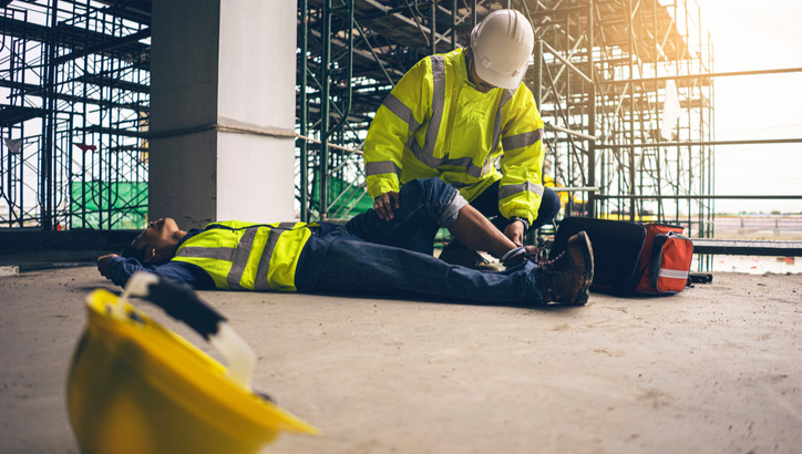 A construction worker checks on a colleague lying on the ground, appearing to give first aid, with scaffolding and a hard hat nearby.