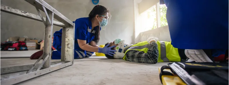 A medical worker wearing a mask assists an injured person in a safety vest lying on the floor inside a building.