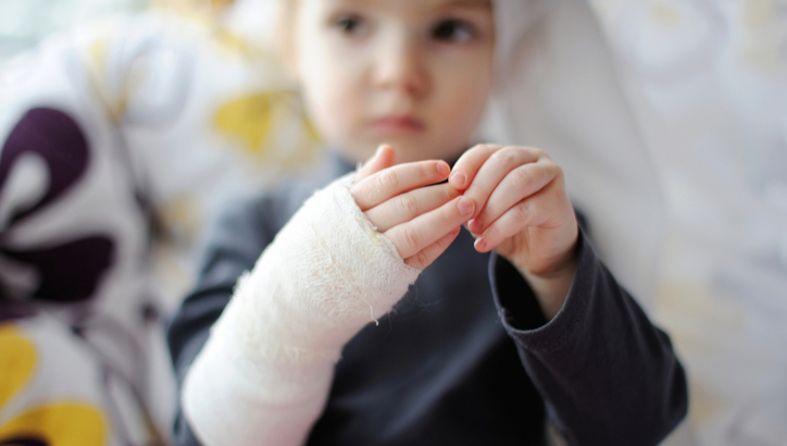 A child with a bandaged hand sits on a patterned pillow, looking away from the camera and touching their injured hand.