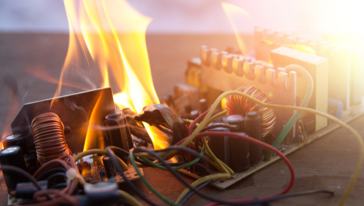 A circuit board is on fire, with visible flames and smoke rising from damaged electronic components and wires.