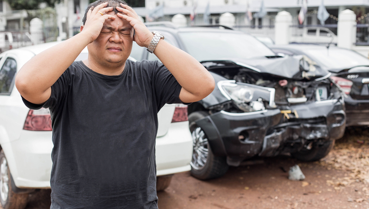 A man stands holding his head in distress in front of a damaged black car involved in an apparent accident.