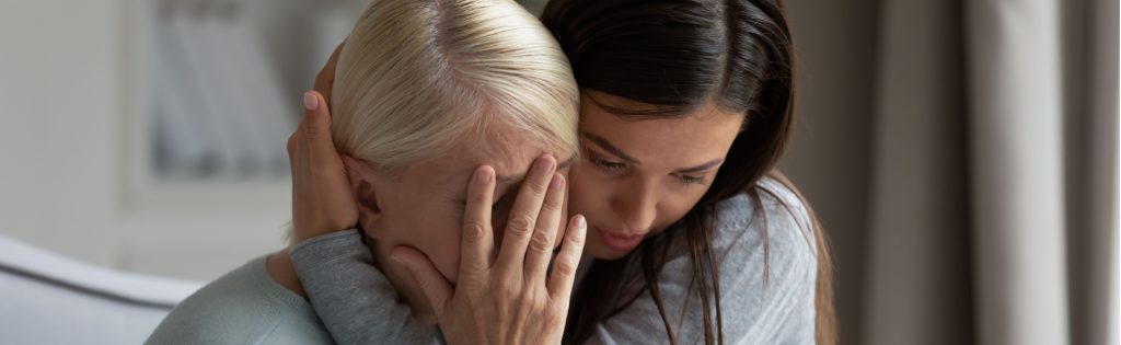 A young woman hugs and comforts an older woman who is covering her face with her hand, both seated indoors.