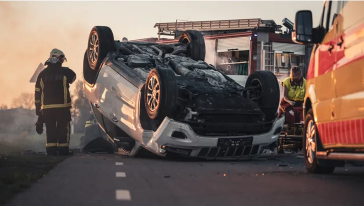 Firefighters and emergency responders attend to a scene where a white car is overturned on a road, with fire trucks and other emergency vehicles present.