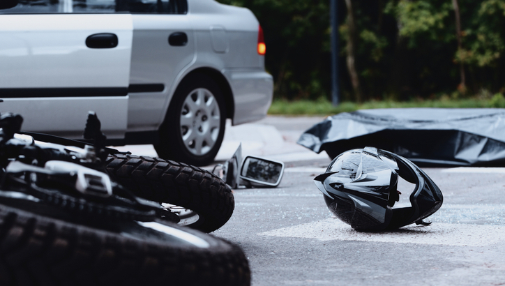 A motorcycle, helmet, and side mirror lie on the road next to a car and a covered body, suggesting a serious accident scene.