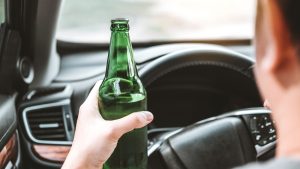 A person holding a green bottle while sitting in the driver’s seat of a car, with their hand on the steering wheel.