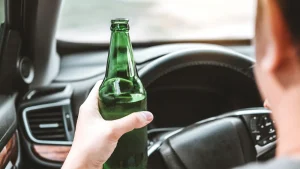 A person holding a green bottle while sitting in the driverÔÇÖs seat of a car, with their hand on the steering wheel.