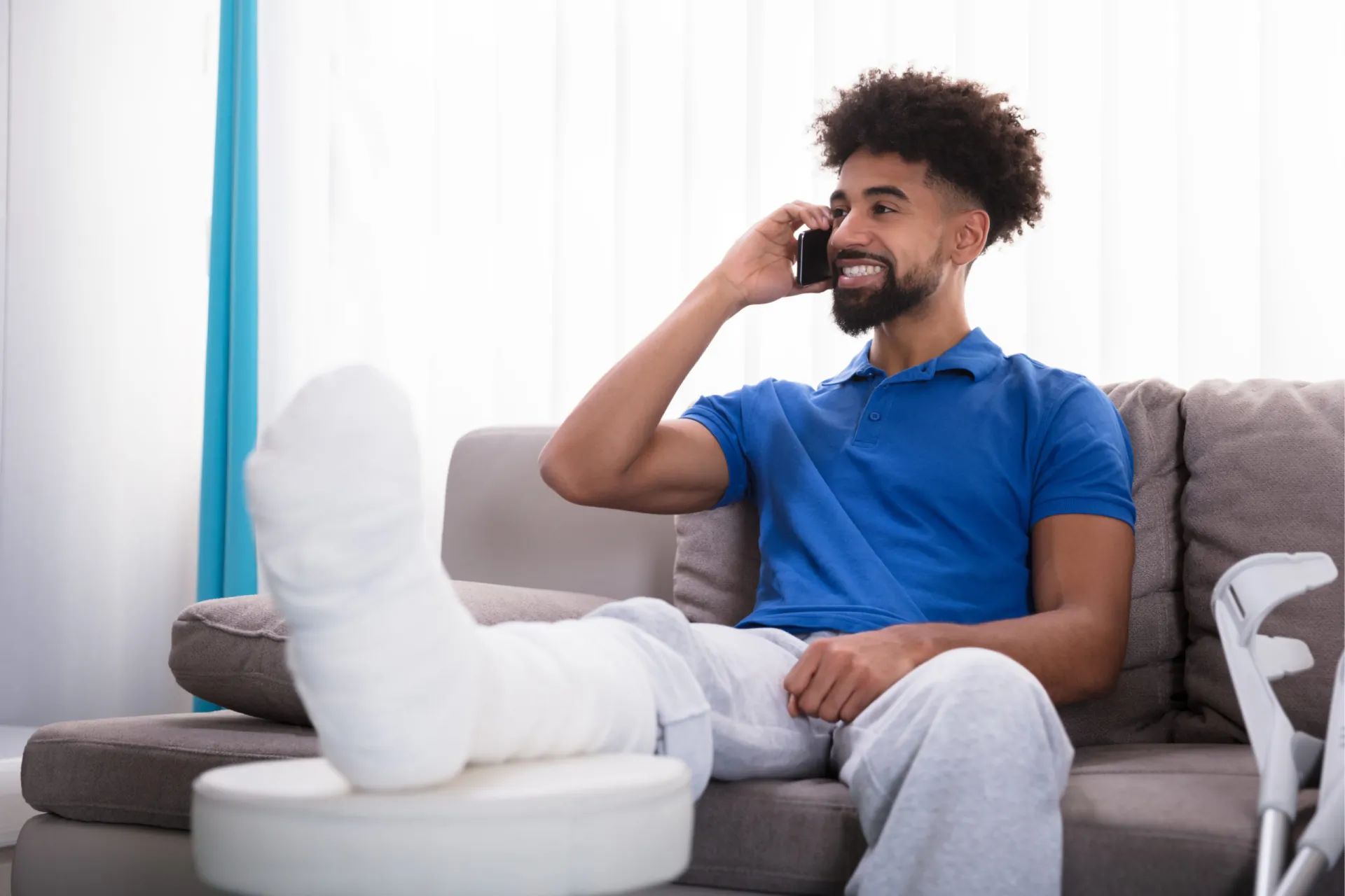 A man with a leg cast sits on a couch, smiling and talking on the phone, with crutches beside him.