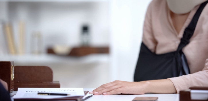 A person with an arm in a sling sits at a desk with paperwork and a smartphone in front of them.