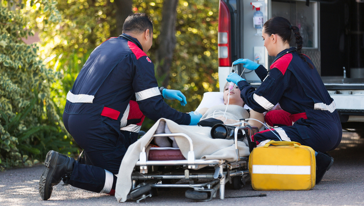 Two paramedics attend to a patient on a stretcher outside an ambulance, providing medical care and using oxygen equipment.