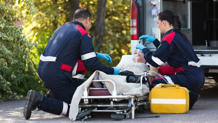Two paramedics attend to a patient on a stretcher outside an ambulance, providing medical care and using oxygen equipment.