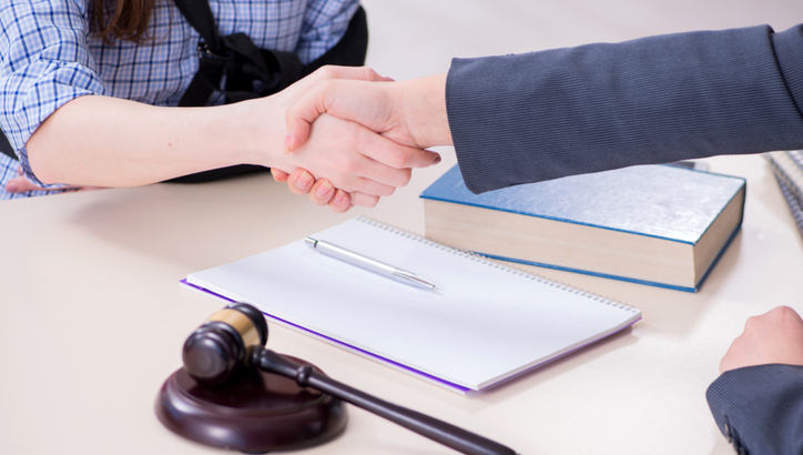 Two people shake hands across a desk with a notepad, pen, gavel, and a book, indicating a legal or professional agreement.