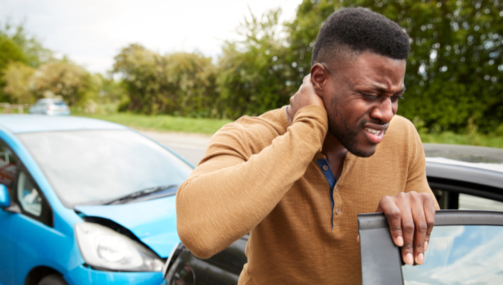 A man stands by a car holding his neck in pain after a rear-end collision with a blue vehicle on a road.