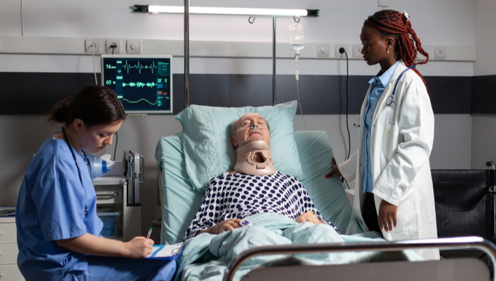 An elderly patient with a neck brace lies in a hospital bed, while a nurse takes notes and a doctor stands by the bedside. Monitors and medical equipment are visible.