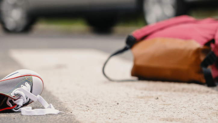 A sneaker with untied laces and a backpack lie on a crosswalk, with blurred vehicles in the background.
