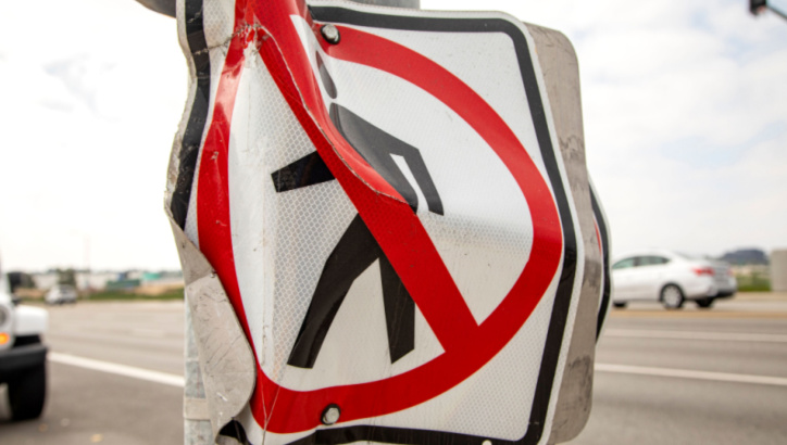 A bent and damaged "No Pedestrian Crossing" sign is attached to a post beside a road with cars in the background.
