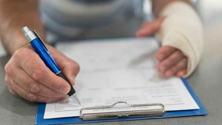 A person with a bandaged hand fills out a medical history form on a clipboard using a pen.