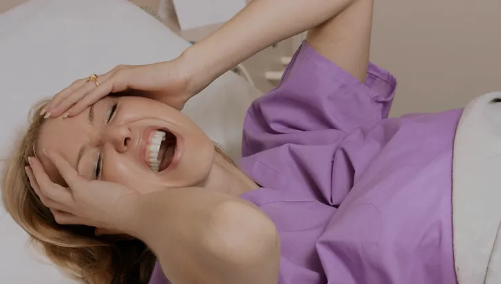 A woman in a purple gown lies in a hospital bed, holding her head and appearing to be in pain or distress.