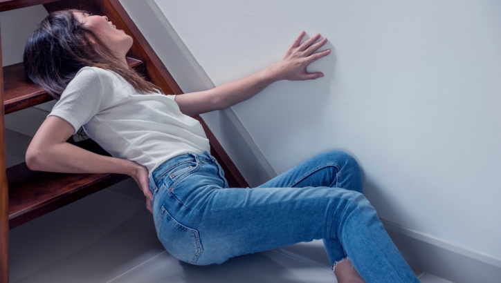 A woman sits on the floor at the base of stairs, holding her lower back and leaning against the wall, appearing to be in pain.