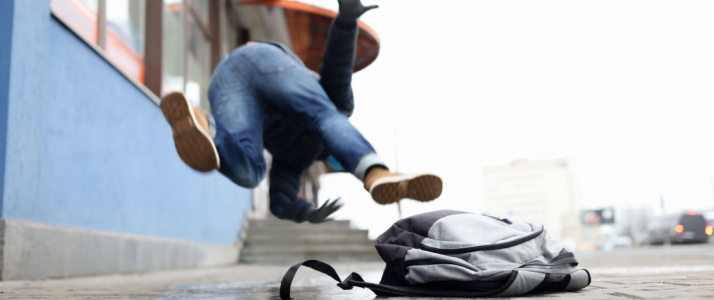 Person falling face-first onto a sidewalk with arms outstretched, while a gray backpack lies on the ground in front of them.