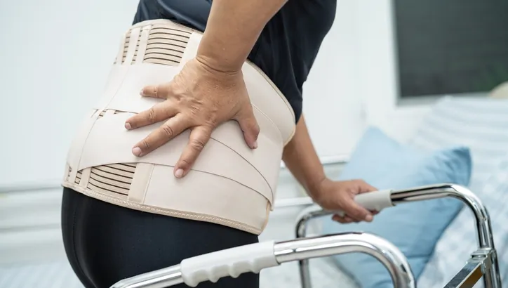 A person wearing a back brace places one hand on their lower back and holds a walker with the other hand, appearing to be in discomfort.