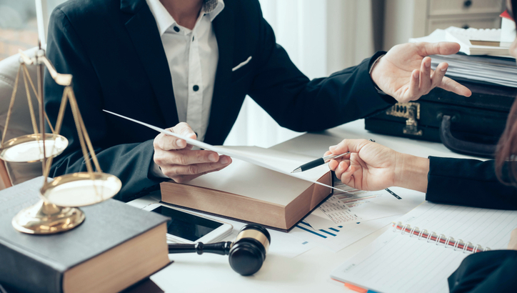 Two people in business attire discuss documents at a desk with legal books, a gavel, scales of justice, a tablet, and papers.