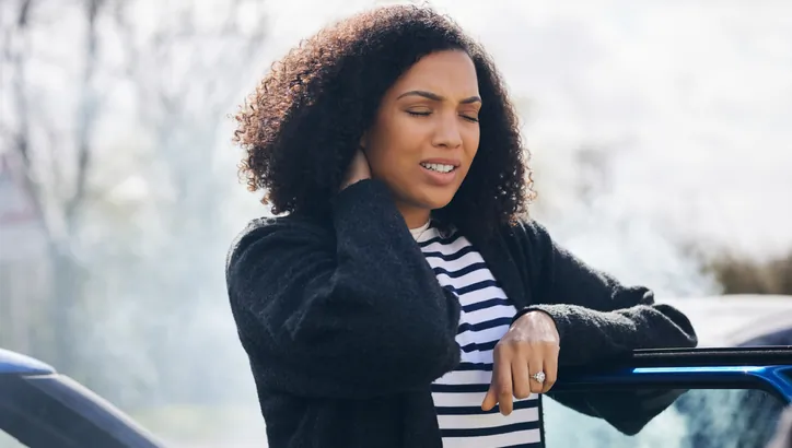 A woman stands outside a car with her eyes closed and one hand on her neck, appearing to be in pain or discomfort. Smoke is visible in the background.