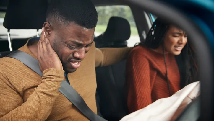 Two people sit in a car after an accident, both appearing in pain; the man holds his neck and the airbags have deployed.