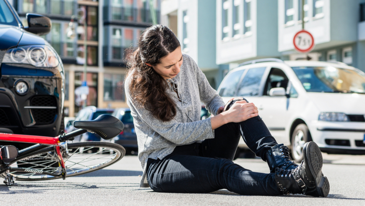 A woman sits on the street holding her injured knee next to a fallen bicycle, with cars parked nearby.