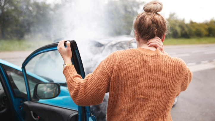 A person stands beside an open car door on the roadside, looking at a crashed vehicle with smoke rising from its hood.