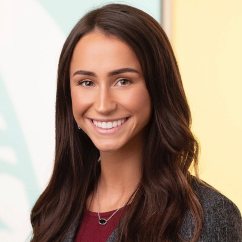 A woman with long brown hair, wearing a dark blazer and a necklace, smiles at the camera in a brightly lit indoor setting.