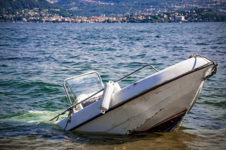 A small white boat is partially submerged and sinking near the shore of a lake, with a distant town visible across the water.